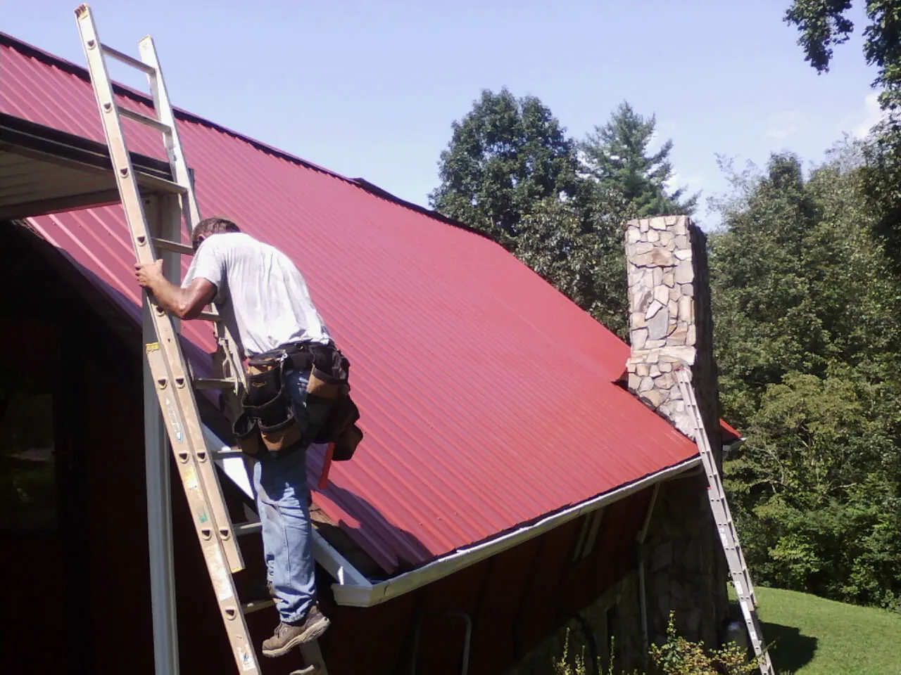 Roofer on a ladder beside a red metal roof and stone chimney — Soffit & Fascia Repair by Roofing Brothers in Pamplico